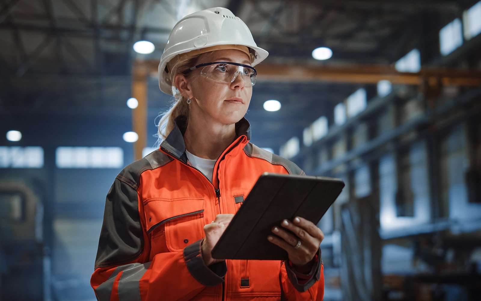 Frau mit Schutzhelm, Brille und Tablet in Werkshalle – Symbolbild für Durchführung der Arbeitsschutzunterweisung